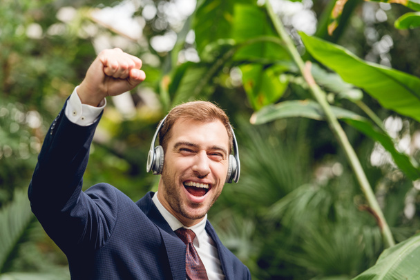 hombre de negocios feliz en traje y auriculares inalámbricos que muestran sí gesto en invernadero
 - Foto, Imagen
