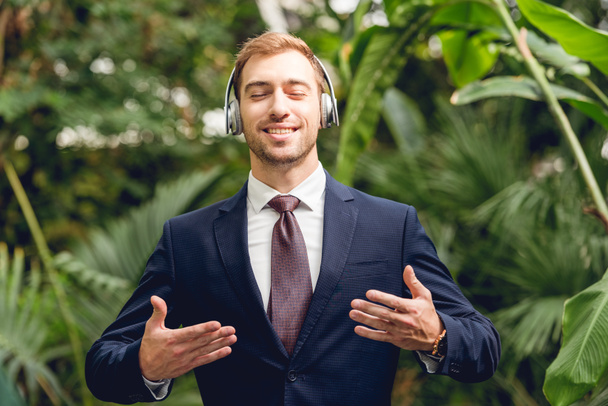 happy businessman in suit and wireless headphones breathing fresh air in greenhouse - Photo, Image