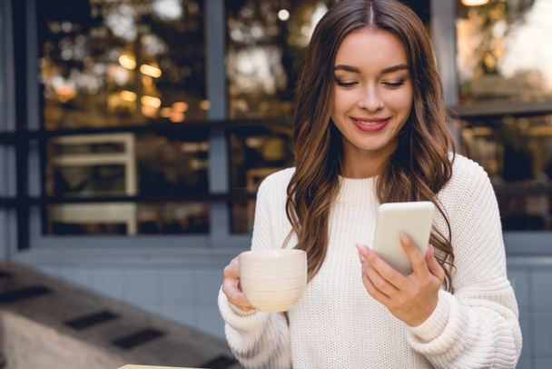 cheerful young woman looking at smartphone while holding cup  - Photo, Image