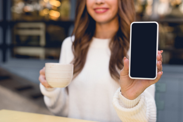 cropped view of cheerful young woman holding smartphone with blank screen and cup  - Photo, Image