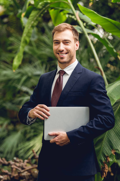 smiling businessman in suit and tie holding laptop in orangery - Fotoğraf, Görsel