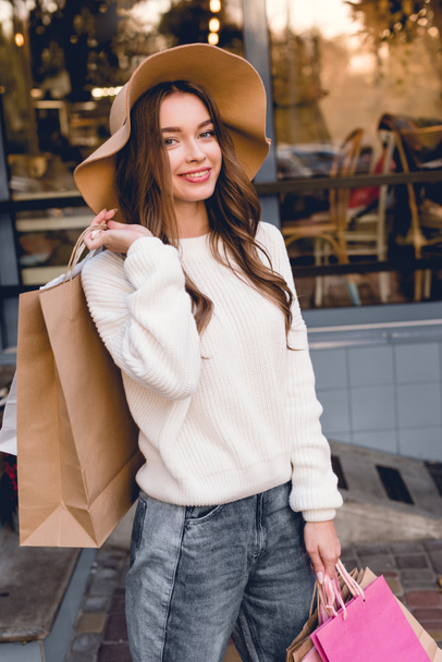 cheerful girl in hat smiling while holding shopping bags  - Zdjęcie, obraz