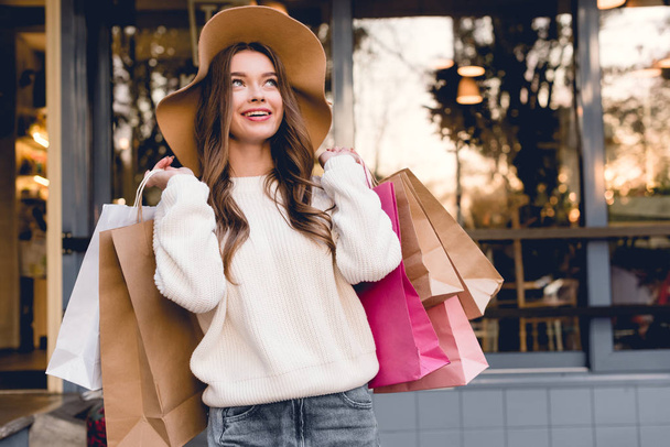 heureuse jeune femme en chapeau souriant tout en tenant des sacs à provisions
  - Photo, image