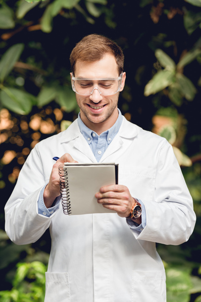 smiling scientist in white coat and goggles making notes in green orangery - Foto, Imagen