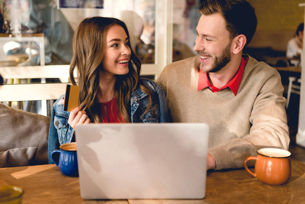 happy young woman holding credit card and looking at cheerful man near laptop  - Фото, изображение