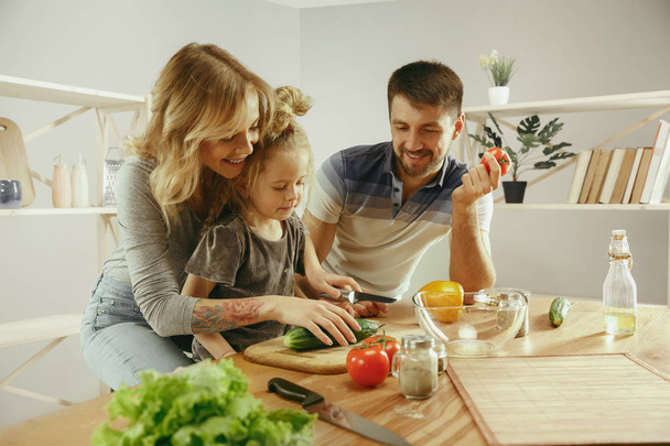 Cute little girl and her beautiful parents are cutting vegetables in kitchen at home - Foto, Imagen