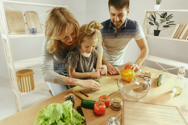 Милая маленькая девочка и ее красивые родители режут овощи на кухне дома
 - Фото, изображение