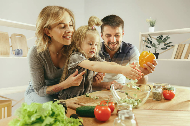 Cute little girl and her beautiful parents are cutting vegetables in kitchen at home - Foto, Bild