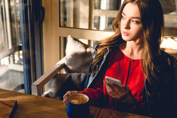 attractive young woman holding cup of coffee and smartphone in cafe  - Photo, Image