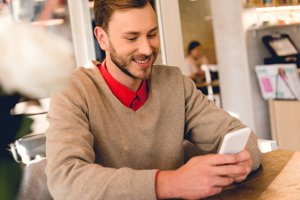 schöner bärtiger Mann schaut auf Smartphone und lächelt im Café  - Foto, Bild