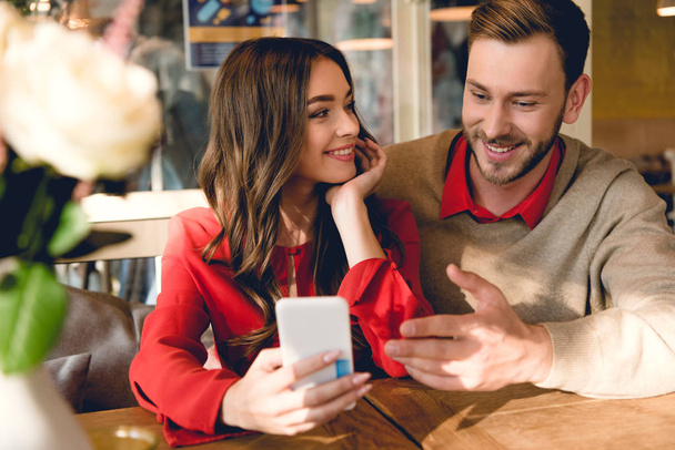 cheerful young woman looking at man gesturing while looking at smartphone  - Photo, Image
