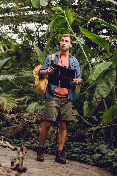 handsome traveler with binoculars and backpack holding map and walking in green tropical forest - Photo, Image