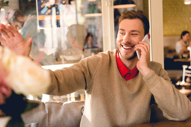 Veselý vousatý muž, který mával rukou a usmíval se při mluvení smartphone v kavárně  - Fotografie, Obrázek