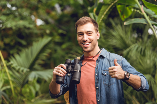 handsome smiling traveler with binoculars and backpack showing thumb up in green tropical forest - Foto, Imagen