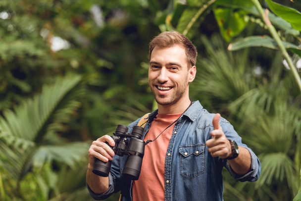 handsome smiling traveler with binoculars and backpack pointing with finger at camera in green tropical forest - Photo, Image