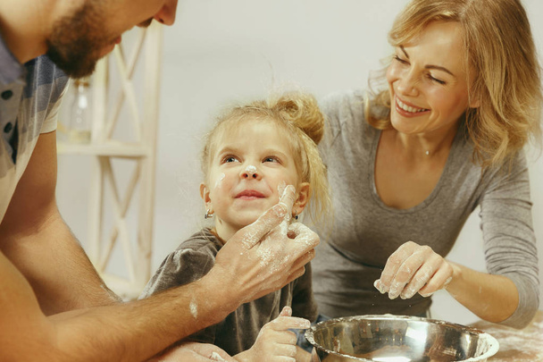 süßes kleines Mädchen und ihre schönen Eltern bereiten den Teig für den Kuchen zu Hause in der Küche zu - Foto, Bild