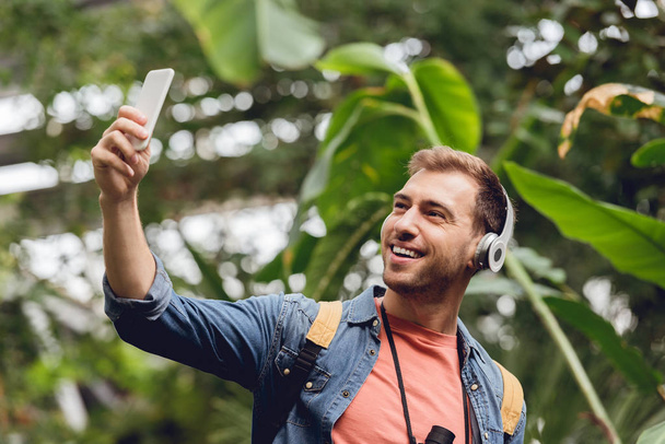 happy traveler in headphones taking selfie in green tropical forest - Foto, Bild