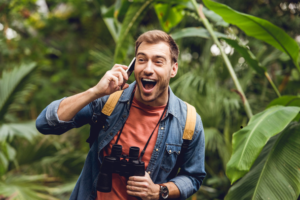 happy traveler with backpack and binoculars talking on smartphone in green tropical forest - Foto, Imagem