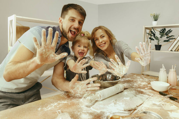 Cute little girl and her beautiful parents preparing the dough for the cake in kitchen at home - Zdjęcie, obraz