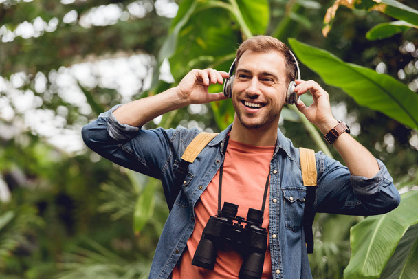 Glücklicher Reisender mit Rucksack und Fernglas, der im tropischen Wald mit Kopfhörern Musik hört - Foto, Bild