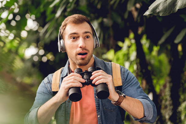 surprised traveler with backpack and binoculars listening music in headphones in green tropical forest - Photo, Image