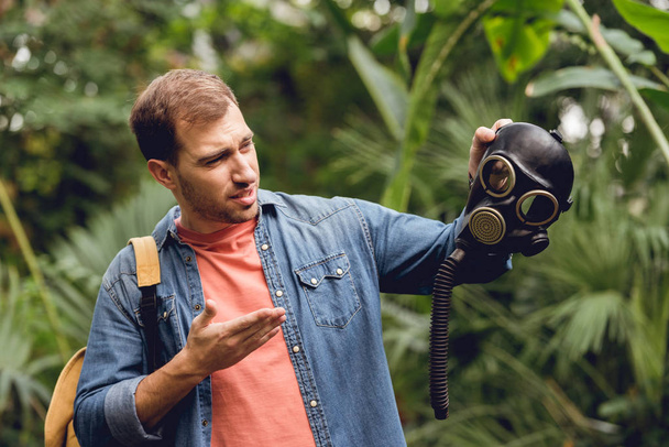 confused traveler with backpack pointing at gas mask in tropical forest - Foto, Imagen