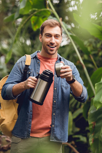 selective focus of happy traveler with backpack opening thermos in tropical forest - Foto, Imagen