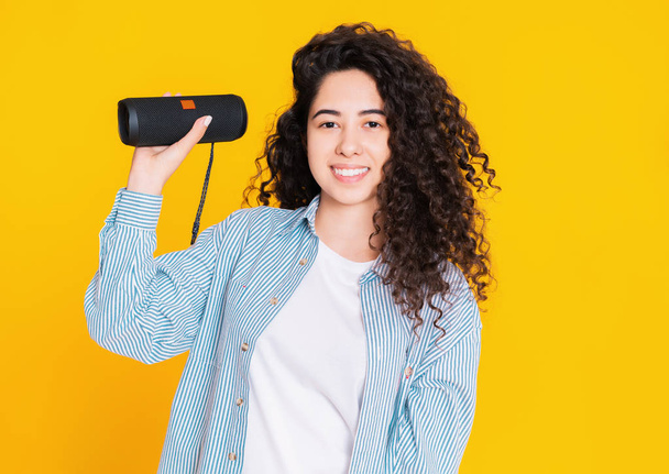 Modern trendy girl listening to music by wireless portable speaker. Young beautiful woman with curly hairstyle enjoying and dancing at yellow background. Female moves to the rhythm of music - Foto, immagini