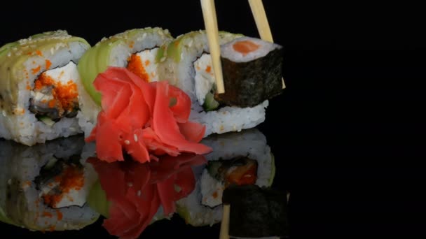 Wooden bamboo Chinese chopsticks put colored sushi rolls and ginger on a mirror surface against a black background. Japanese cuisine in studio - Footage, Video