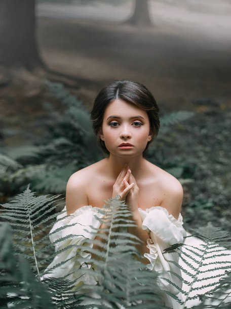 small newborn wood elf is begging for mercy, the spirit as a symbol of fear and danger before cruel people, cute dark-haired girl sits in tropical green leaves, dark forest in the glare of sunlight - Foto, Bild