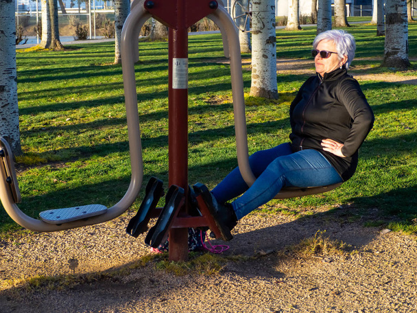 Μια ηλικιωμένη γυναίκα με άσπρα μαλλιά που εξασκώ τη γυμναστική σε ένα βιο-υγιές πάρκο - Φωτογραφία, εικόνα
