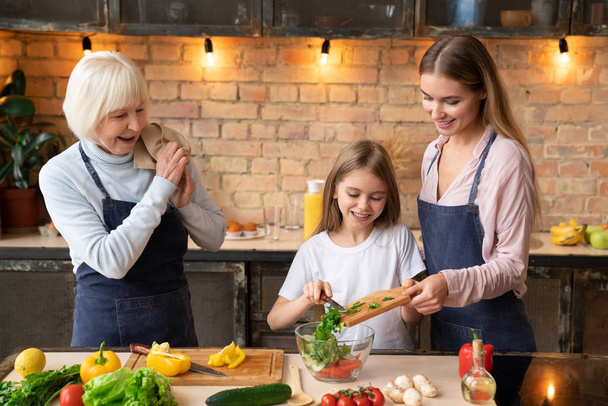 Beautiful woman helping her little daughter to prepare ingredients for fresh salad. Grandmother watching with towel in hand - Foto, Imagen