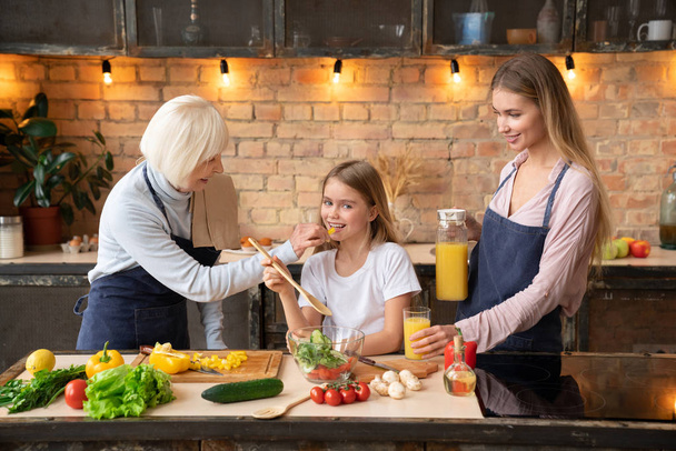 Old lady feeding her granddaughter with pepper while cooking salad at home. Smiling mother standing with glass of juice in her hand on the kitchen table - Photo, image