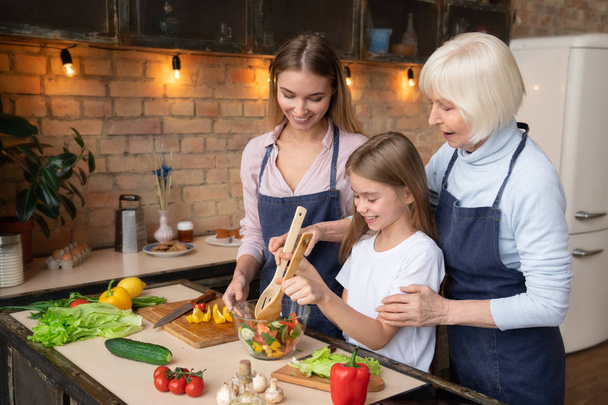 Side view shot of grandmother teaching granddaughter to mix salad with wooden spoons in glass bowl. Smiling mother looking on them in kitchen - Photo, image