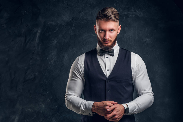 Elegantly dressed bearded hipster in a vest with bow tie. Studio photo against a dark wall background - Fotografie, Obrázek