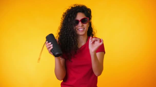 Modern trendy girl listening to music by wireless portable speaker. Young beautiful woman with curly hairstyle enjoying and dancing at yellow background. Female moves to the rhythm of music. - Πλάνα, βίντεο