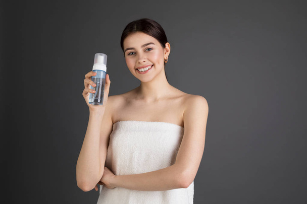 Милая элегантная улыбающаяся женщина с голыми плечами держит и представляет косметическую бутылку с пеной
 - Фото, изображение