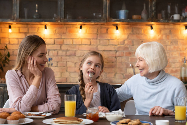 Bonne petite fille qui s'amuse au petit déjeuner avec sa mère et sa grand-mère. Crêpes, gâteaux, jus frais et confiture sur la table
 - Photo, image