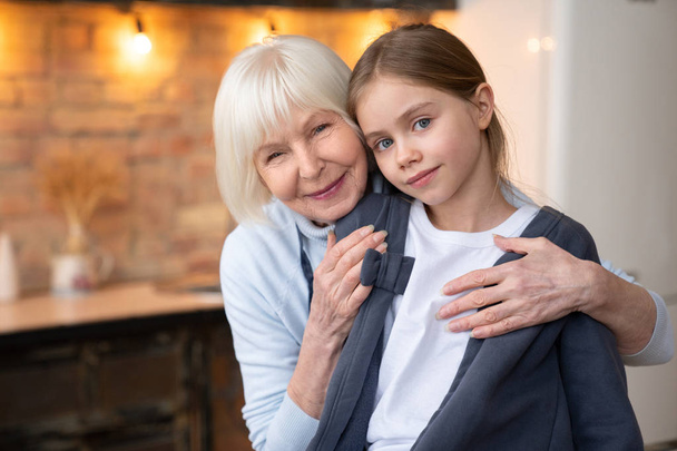Mature grandmother hugging her granddaughter in kithen with look in camera - Valokuva, kuva