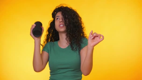 Modern trendy girl listening to music by wireless portable speaker. Young beautiful woman with curly hairstyle enjoying and dancing at yellow background. Female moves to the rhythm of music. - Video