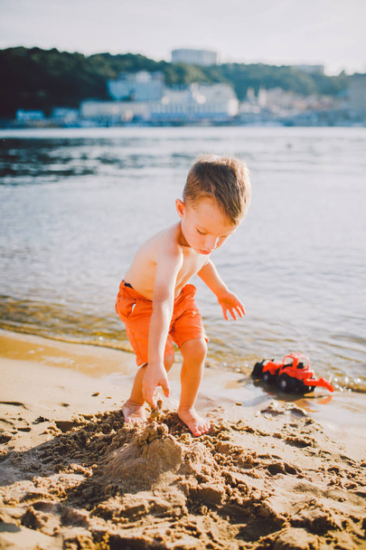 Thema Bau und Schwerindustrie. Abstraktion Junge spielt auf dem Sand in der Nähe des Flusses im Sommer Spielzeug roten Traktor Modell, Baggermaschine mit Eimer - Foto, Bild