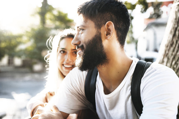 Крупным планом портрет уверенного бородатого человека, смотрящего в сторону улыбающегося, в то время как его обнимает его девушка, которая смотрит на него на улице во время свиданий в городе
. - Фото, изображение