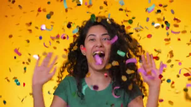 Young latin girl with curly hair dancing and having fun in confetti rain on yellow background. Woman celebrating, depicts joy and happiness. Success, victory, holiday concept. - Video