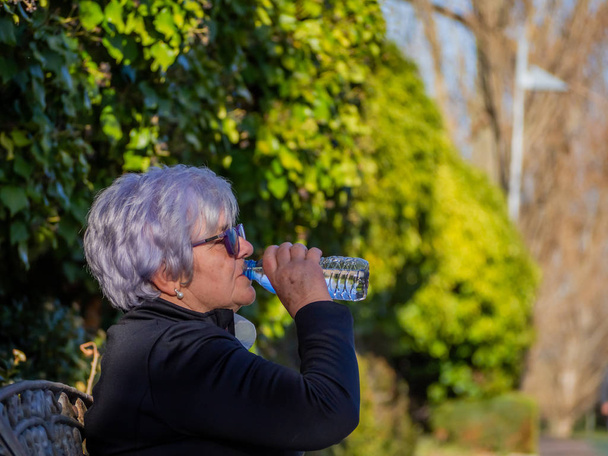 Літня жінка з білим волоссям сидить на лавці парку питної води з пластикової пляшки
 - Фото, зображення