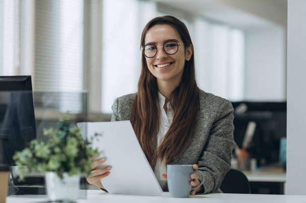 Retrato de una joven y sonriente mujer de negocios con gafas sentada en el lugar de trabajo y trabajando con documentos
. - Foto, imagen