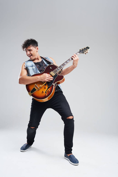 Young man musician with an electric guitar in hand on a gray background. he plays rock and roll loudly. Full-length portrait. - Photo, Image