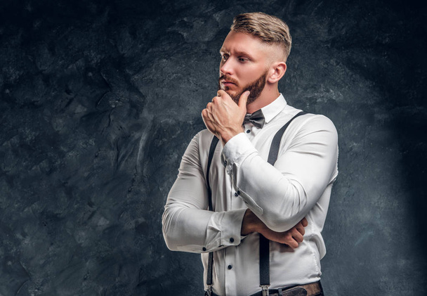 Pensive male thinking about something important. Stylishly dressed young man in shirt with bow tie and suspenders posing with hand on chin. - Photo, image