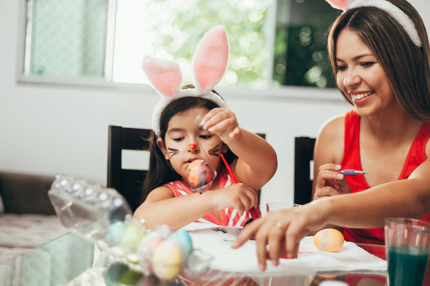 Veselé velikonoce! Matka a její dcera, malování kraslic. Šťastná rodina, příprava na Velikonoce. Roztomilé dítě holčička nosit uši na velikonoční den - Fotografie, Obrázek