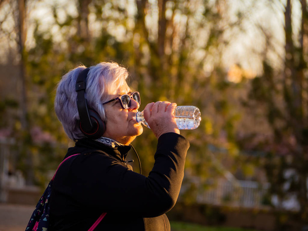 Μια ηλικιωμένη γυναίκα με άσπρα μαλλιά ακούγοντας μουσική με ακουστικά και πόσιμο νερό από ένα πλαστικό μπουκάλι - Φωτογραφία, εικόνα