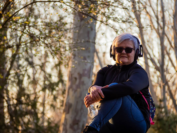 Uma mulher idosa com cabelos brancos ouvindo música com fones de ouvido e uma garrafa de água de plástico na mão
 - Foto, Imagem